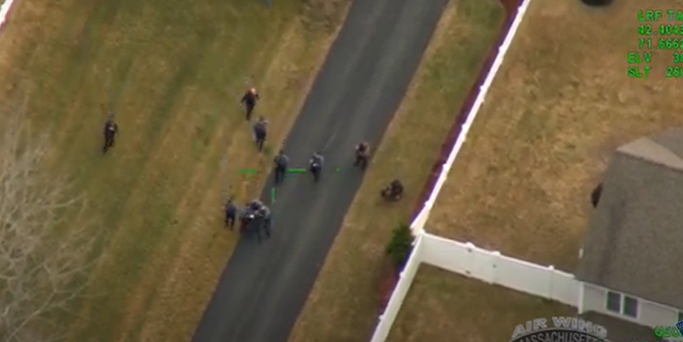 Aerial view of police officers surrounding a suspect on a paved driveway in a suburban neighborhood. Several officers are standing while others are restraining the individual on the ground. A white fence borders a house nearby. The image appears to be from a Massachusetts State Police Air Wing camera, with green targeting indicators and flight data overlayed on the screen.