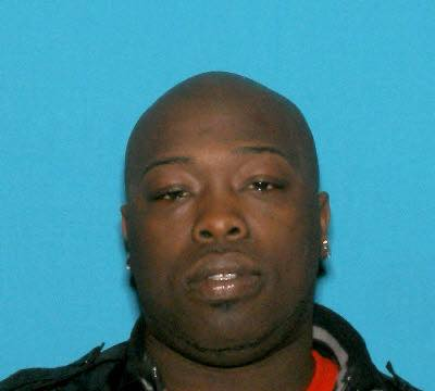 A booking-style photo of a bald Black man with a serious expression, wearing a black jacket over a red shirt. The background is a solid blue color.