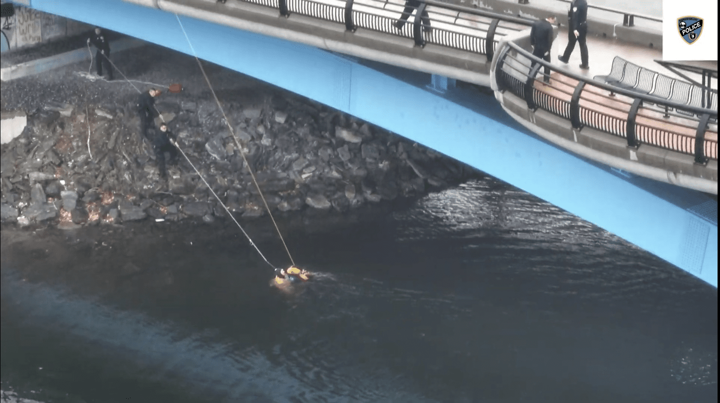 Police officers conduct a water rescue under a blue bridge, using ropes to pull a person in a yellow flotation device toward the rocky shore. Other officers stand on the bridge and embankment assisting in the operation. (Photo credit: Shrewsbury Police Department)