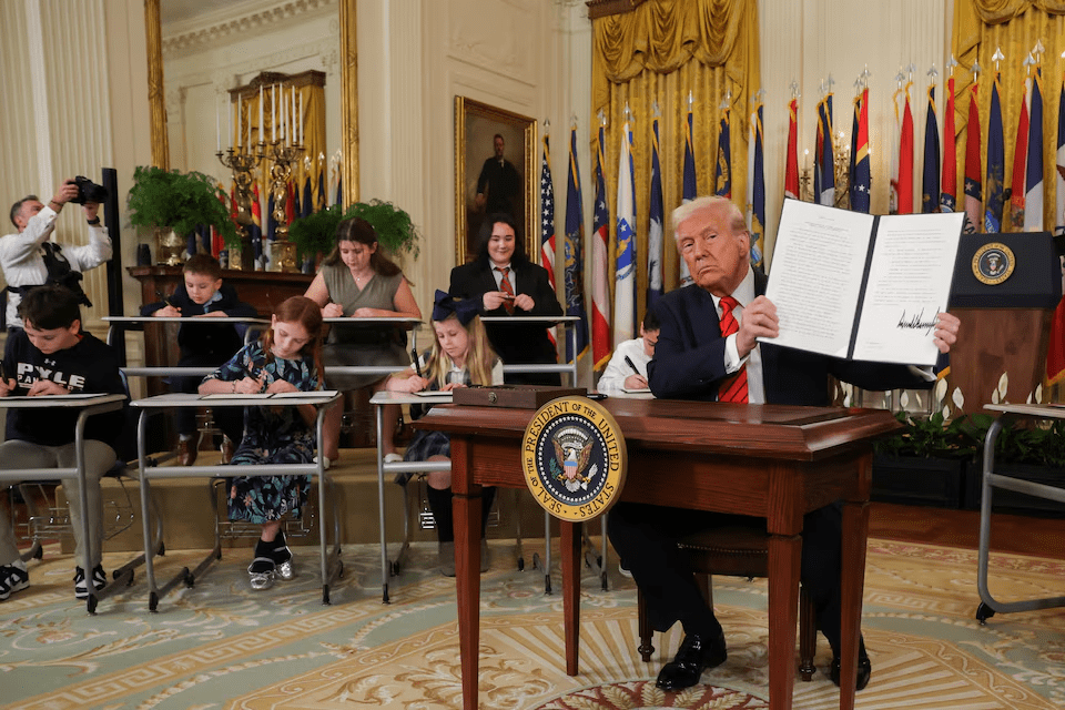 President Donald Trump holds up a signed executive order in the White House, with schoolchildren seated at desks behind him, representing the administration’s push to return educational control to states.