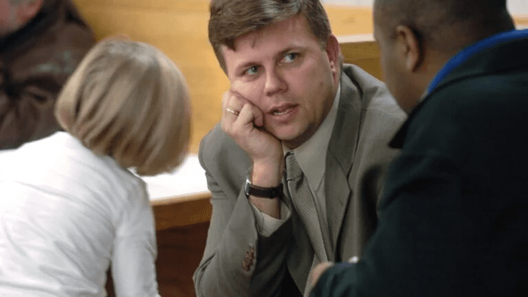 A man in a suit leans on his hand while engaged in conversation with another person in a courtroom. A young girl with blonde hair is seen in the foreground. The setting appears to be a public hearing or meeting.