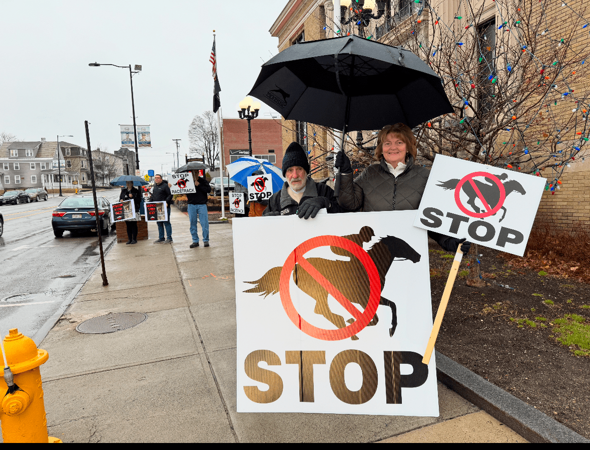 Protesters stand on a rainy sidewalk outside Leominster City Hall holding signs with horse racing graphics crossed out and the word “STOP,” opposing the proposed Fairgrounds racetrack project.