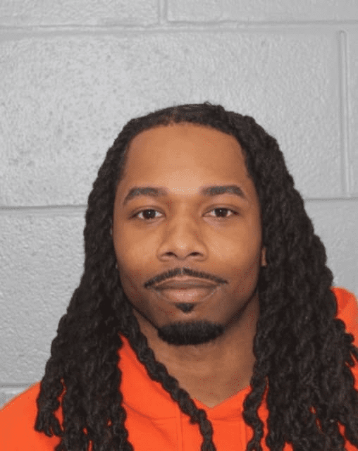 Mugshot of Bilal Griffith, a man with long braids wearing an orange sweatshirt, standing in front of a gray cinderblock wall.