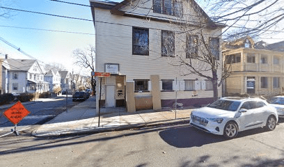 A street view of a white, three-story building on a corner lot with a sign reading "Central Street Studios." A white car is parked on the road, and a bright orange "Sidewalk Closed Ahead" sign is posted nearby. Trees without leaves and surrounding residential homes suggest it is late fall or winter.