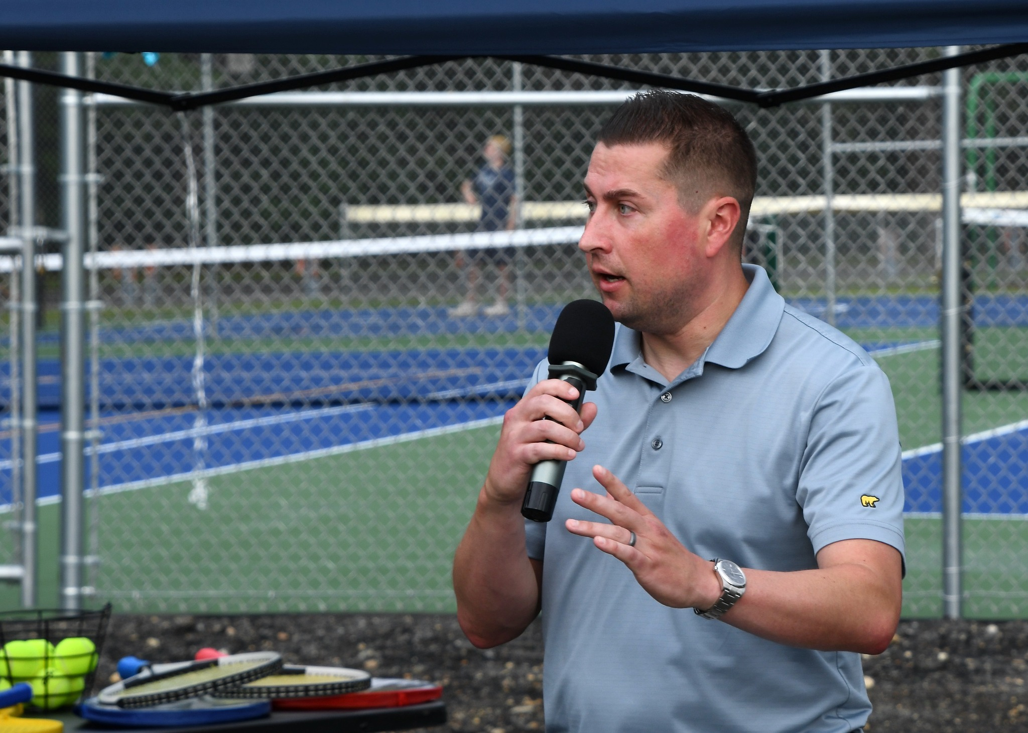 Templeton Town Administrator Adam Lamontagne speaks into a microphone while wearing a light blue polo shirt. He gestures with his left hand while addressing an audience at an outdoor event near a fenced-in sports court with tennis and pickleball equipment in the foreground.