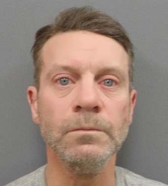 A mugshot of a middle-aged man with short brown hair, light stubble, and blue eyes, wearing a gray shirt. He has a neutral facial expression and is looking directly at the camera against a plain gray background.
