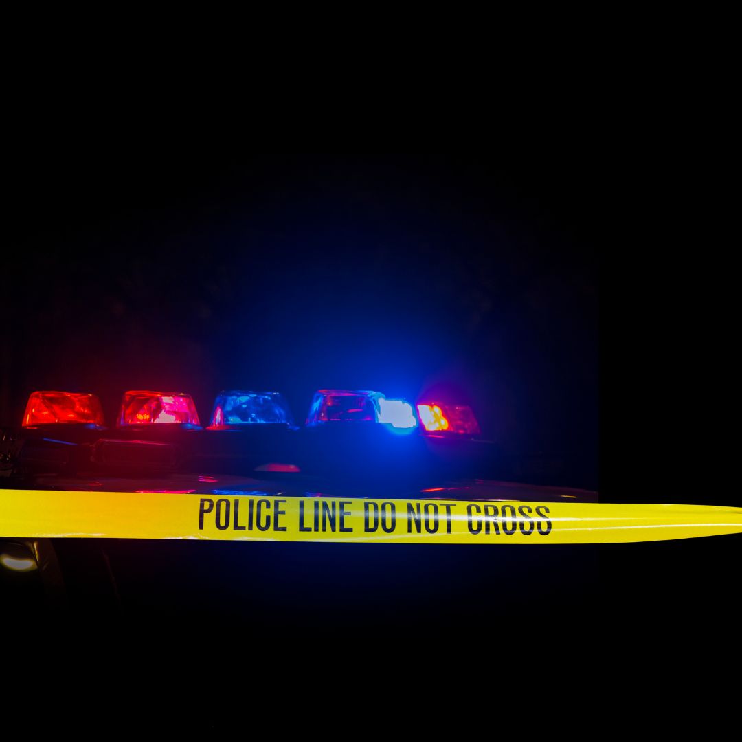 A police vehicle with flashing red and blue emergency lights is seen behind yellow "Police Line Do Not Cross" tape at night, illuminating the dark surroundings.