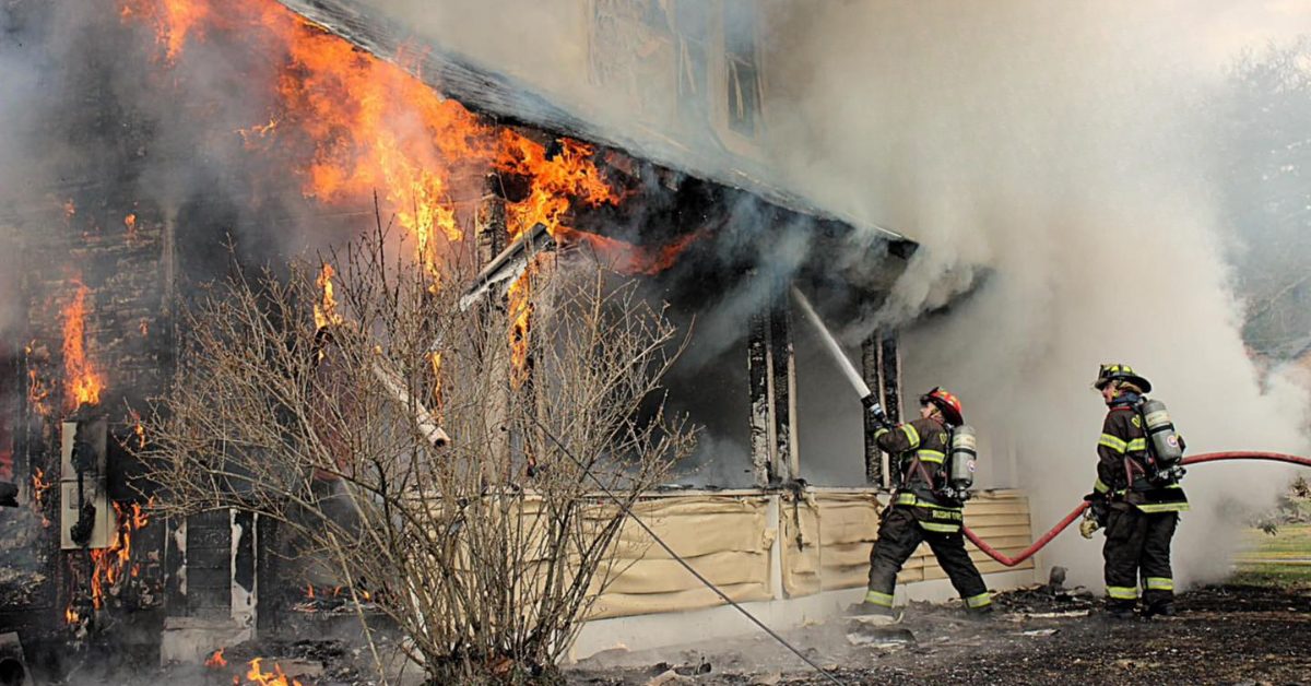 Firefighters spray water on the side of a house engulfed in heavy flames and thick smoke. Photo Credit: Orange Fire Rescue