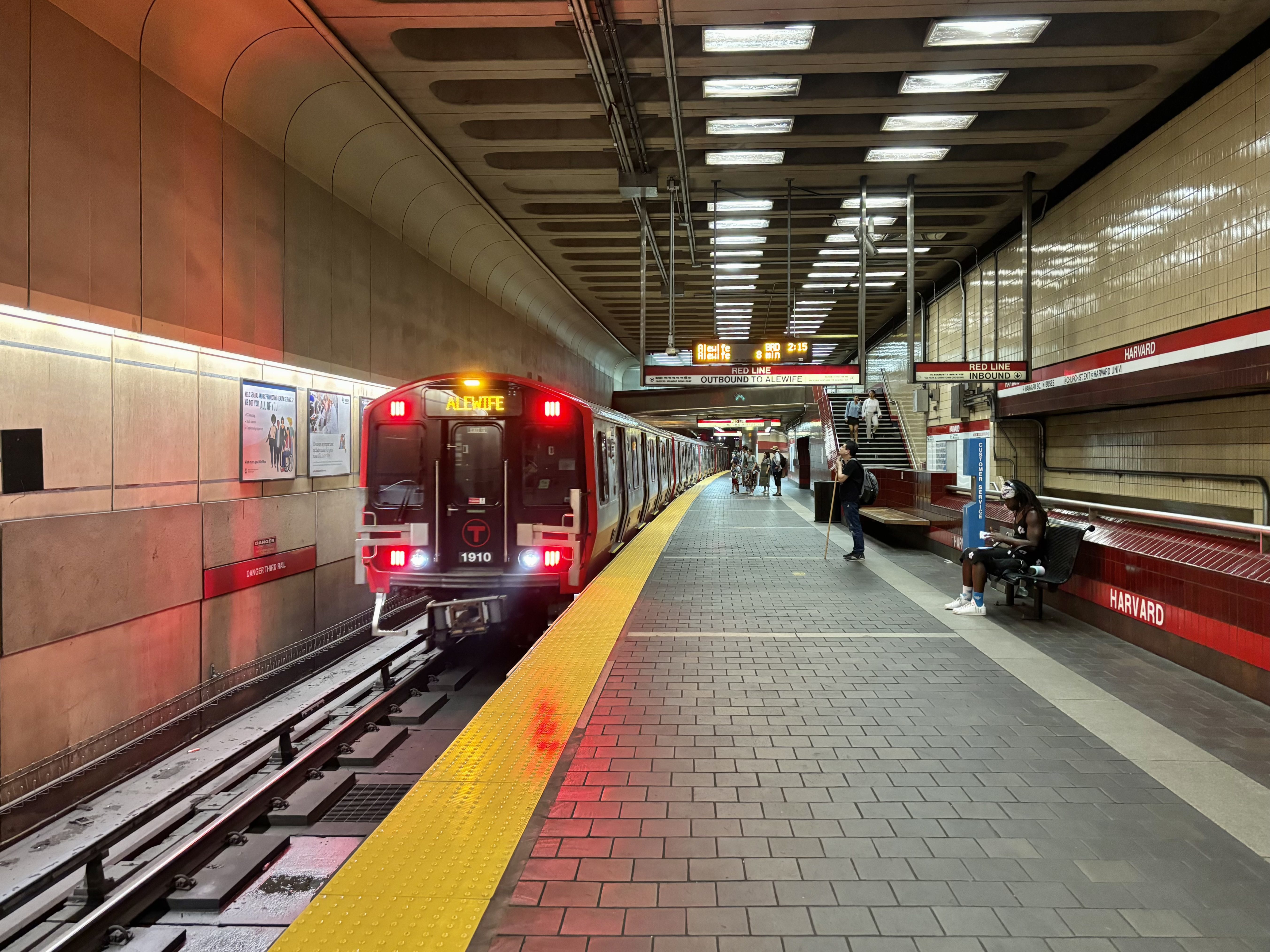 Red Line train arriving at the southbound platform of Harvard Square MBTA station with passengers waiting and signage indicating the outbound direction to Alewife.