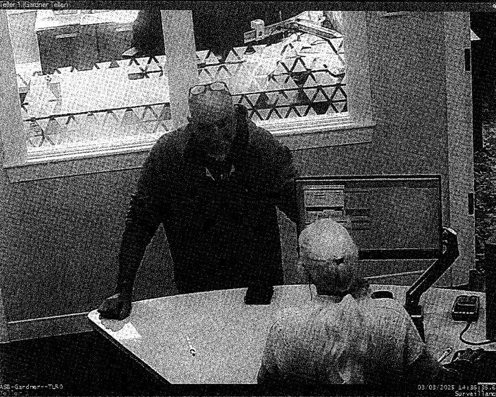Black-and-white surveillance image showing a man standing at a bank counter, interacting with two bank employees. The scene is captured from an overhead angle, with papers and office equipment visible.