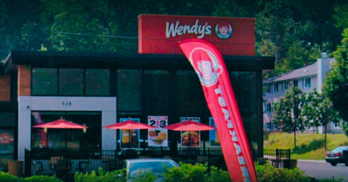 The exterior of a Wendy’s restaurant located at 528 North Main Street in Leominster, Massachusetts, featuring red patio umbrellas, a red Wendy’s feather flag, and promotional posters in the windows.