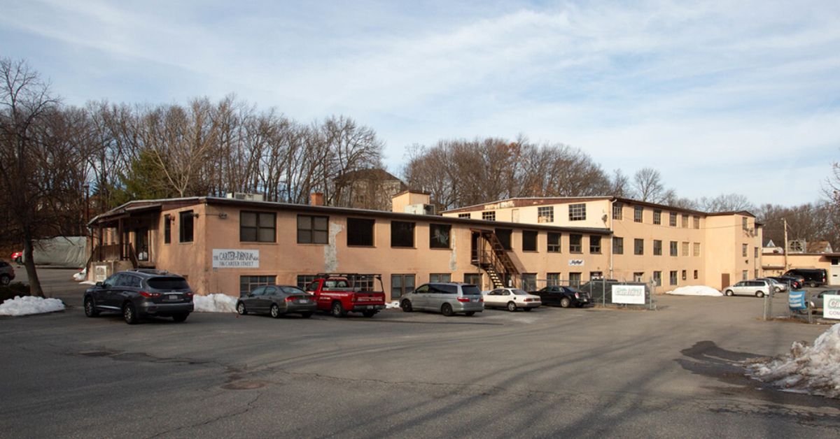 A large beige industrial building at 106 Carter Street in Leominster, surrounded by parked cars, with signs for Carter & Dumas and other businesses on the exterior. Snow piles line the edges of the parking lot.