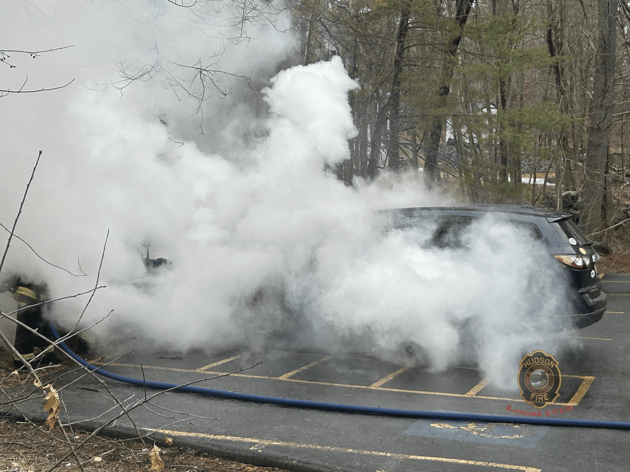 A dense cloud of white smoke envelops a car in a parking lot as firefighters from Hudson Firefighters Local 1713, visible through the smoke, work to control a car fire with a hose extended from their fire truck.