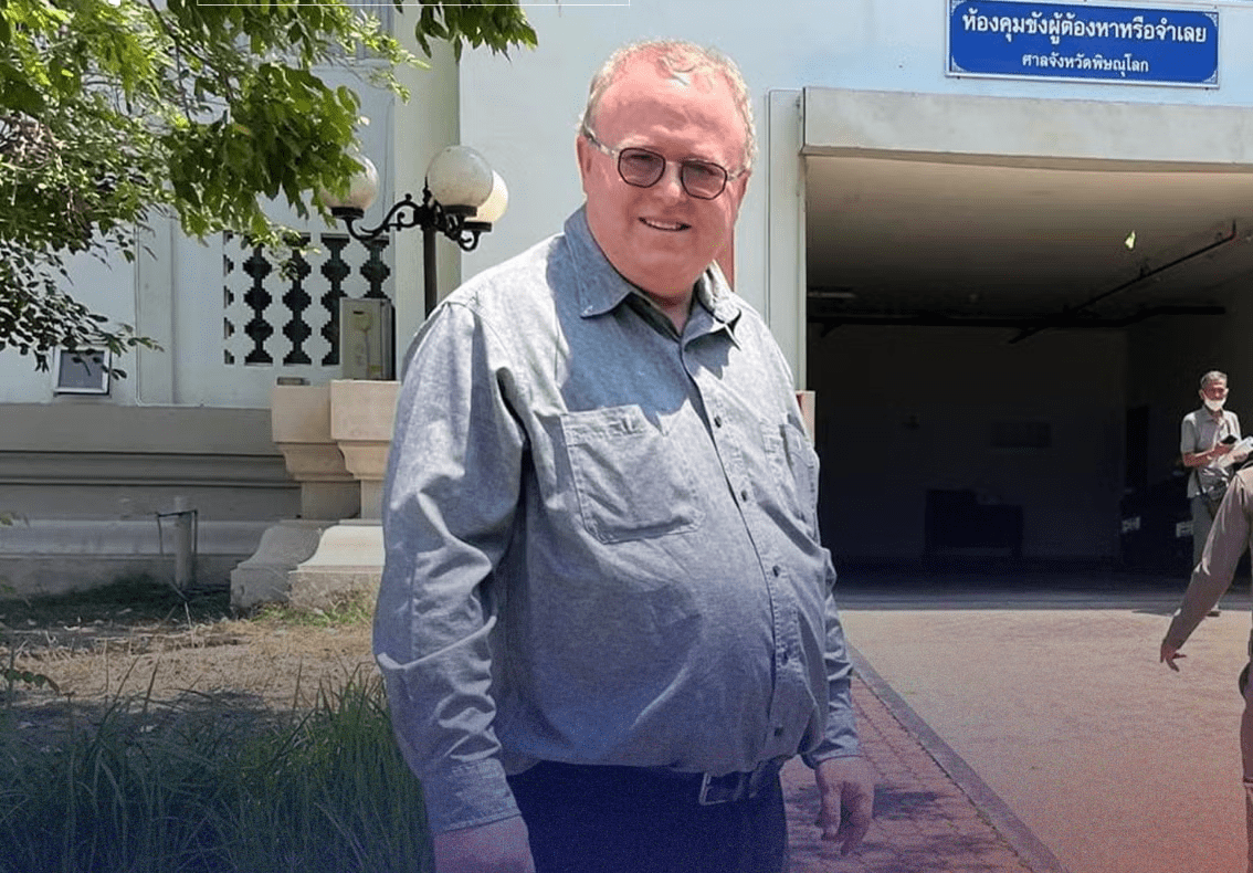 Paul Chambers, an American academic, stands outside a Thai government building escorted by uniformed officers. He wears a face mask and casual clothing while being led by authorities following his arrest.