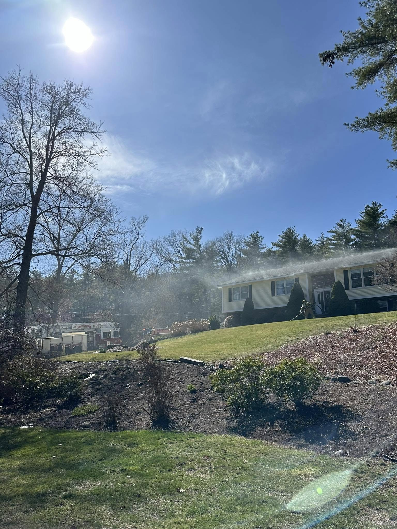 A light-colored split-level home on a hill with smoke rising from the roof, surrounded by trees and a clear blue sky. Firefighters are seen approaching the house.