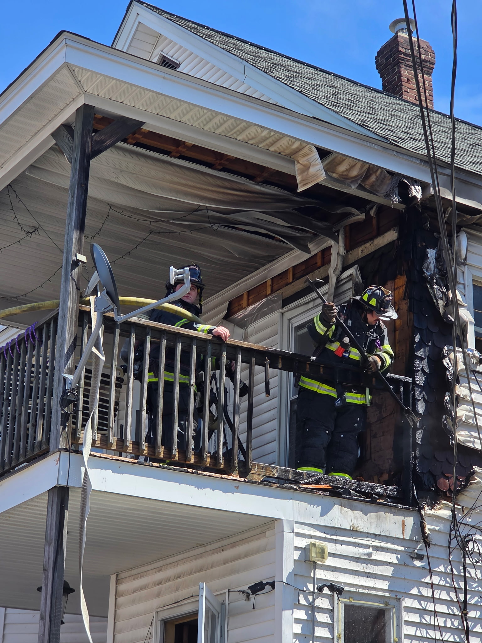 Two firefighters in full gear stand on a second-floor porch of a white home, examining fire damage to the railing and roof following a blaze in South Fitchburg. The siding and soffit are visibly melted, and the firefighters are using tools to check for hot spots.