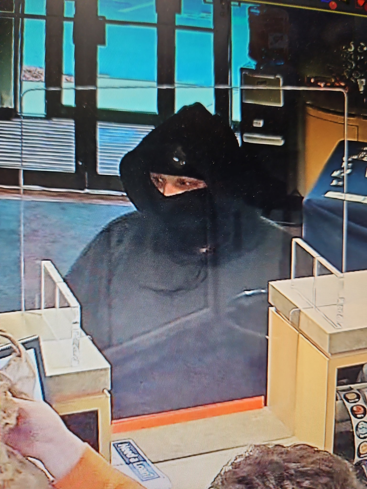 Surveillance photo of a masked suspect inside a credit union wearing a black hooded sweatshirt and black balaclava, with only part of the face visible above the mask.