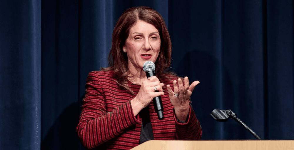 A woman in a red and black striped blazer speaks into a handheld microphone while standing at a podium with a dark blue curtain backdrop. She gestures with her left hand while addressing an audience.