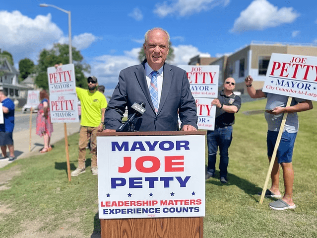 ChatGPT said: Alt text: Mayor Joe Petty stands at a podium outdoors during a campaign event, smiling in a suit and tie, with supporters holding signs that read "Joe Petty for Mayor" in the background.