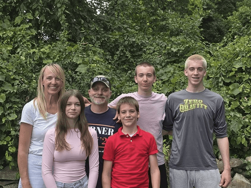 Carolyn Sanger stands with her family outdoors in front of green foliage. She is smiling alongside two adult men and three children, all smiling and casually dressed.