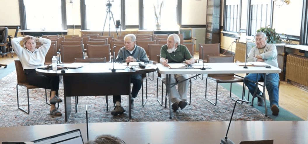 Screenshot of a public-meeting room: four Finance Committee members sit at two tables facing the camera. From left, Erin Powell leans back with hands behind her head, Robert Latini reviews papers, Paul Cherubini speaks while gesturing with a pen, and Bernard Hastings sits with arms folded. Rows of empty audience chairs, large windows and a video tripod appear in the background.