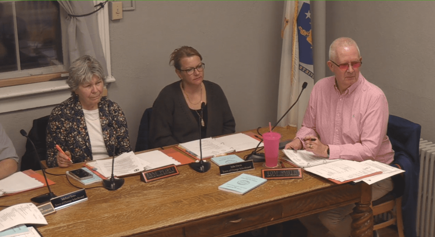 Screenshot of three Orange Select Board members seated at a table during a public meeting. Jan Pierce holds a pen and paper, Julie Davis looks ahead, and Tom Smith wears a pink shirt and red glasses. A Massachusetts state flag stands in the background.