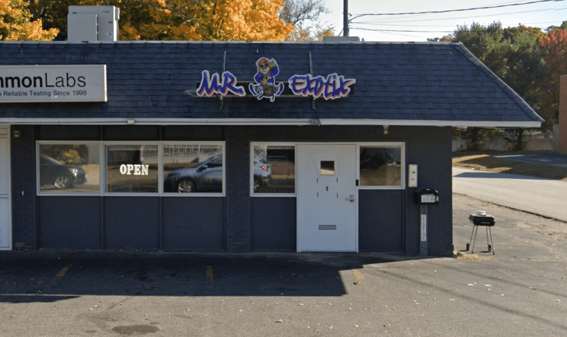 Front view of the Mr. Exotic storefront in Webster, MA, with a dark gray exterior, white-framed windows, an “OPEN” sign in the left window, and a small black grill on the pavement near the entrance.
