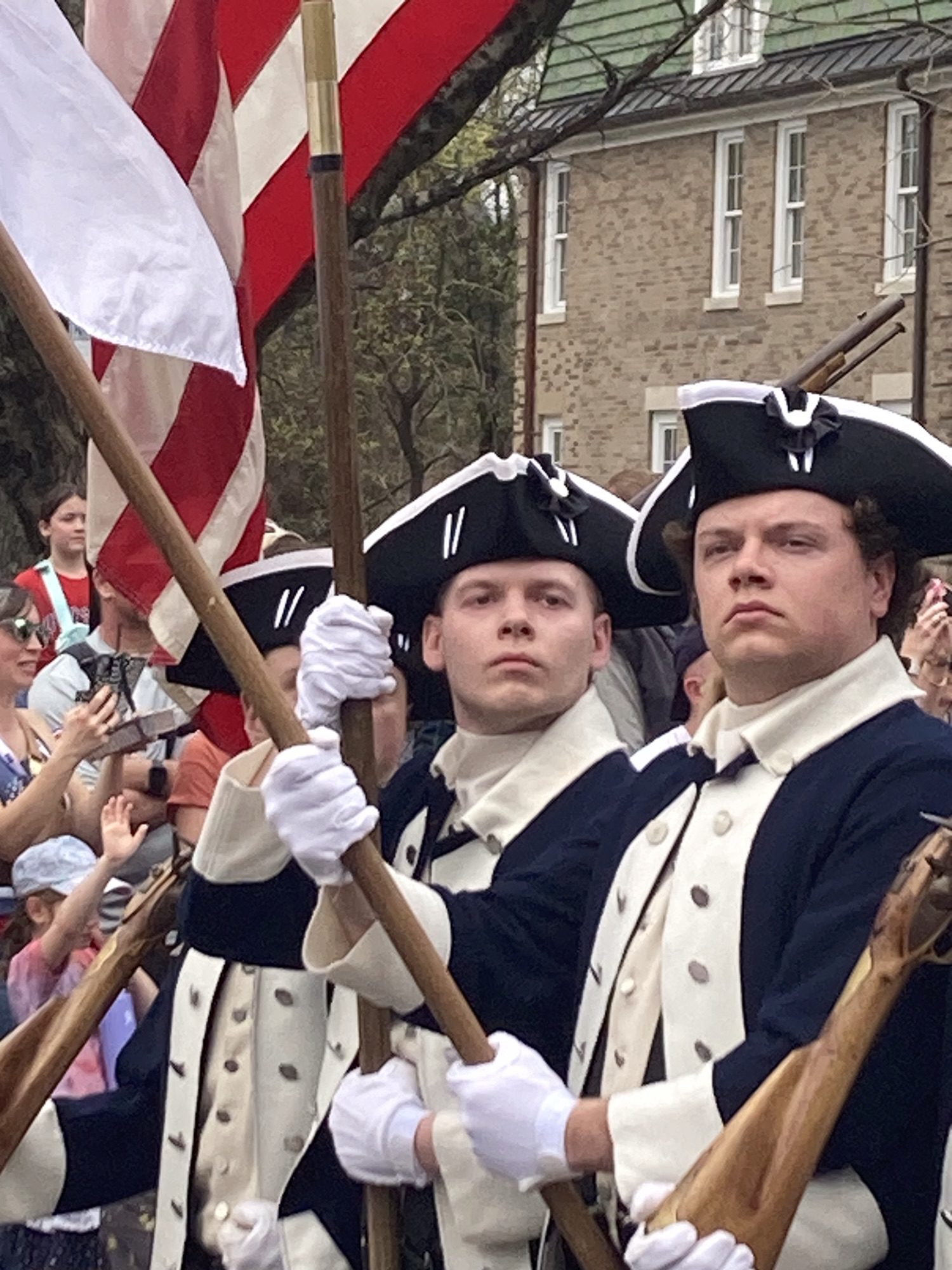 Men dressed in navy and cream colonial-style uniforms hold flags as they march in the 2025 Patriots’ Day Parade in Concord, surrounded by a crowd of onlookers.
