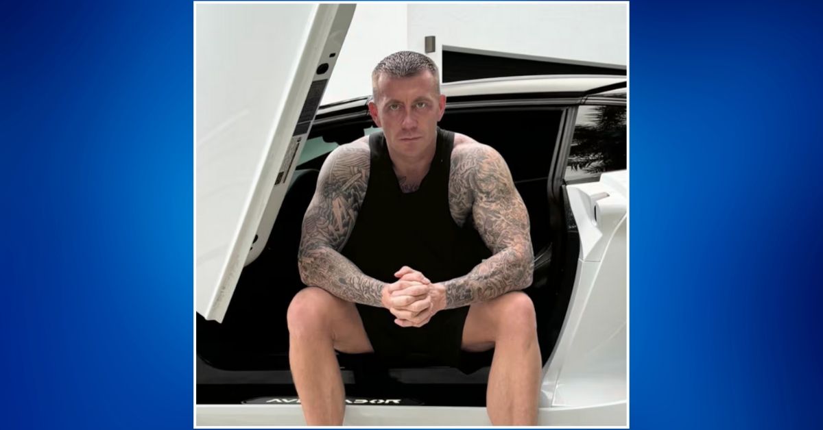 Eric Spofford sits in the open doorframe of a white Lamborghini, wearing a black tank top and shorts, with full tattoo sleeves visible on both arms, staring directly at the camera.