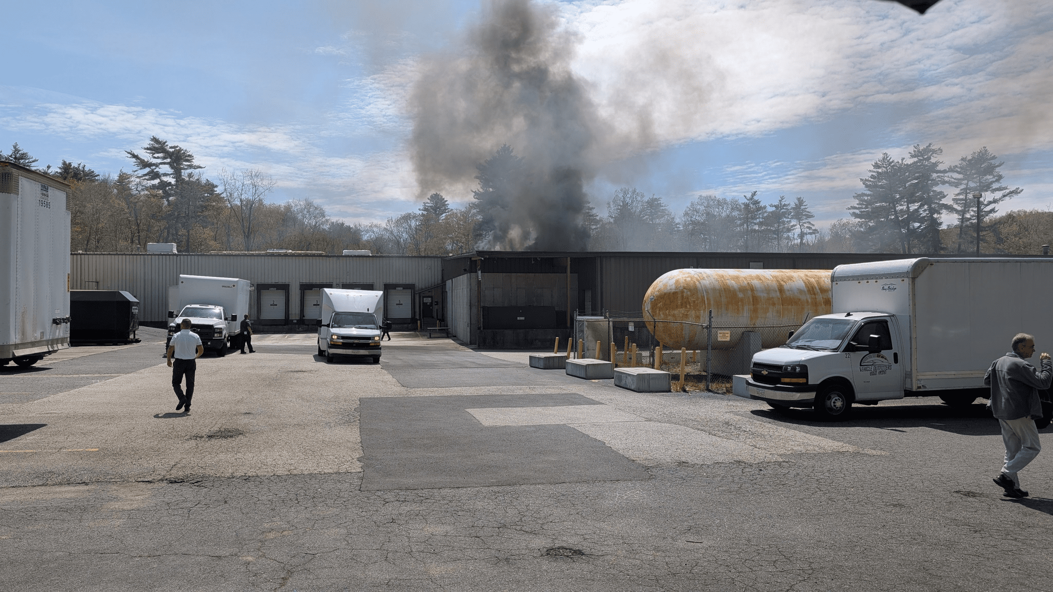A parking lot in front of a large industrial building with black smoke rising from its roof. Several box trucks and emergency responders are visible, and a yellow propane tank sits beside the building.