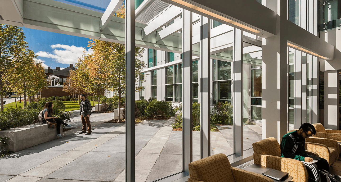 Interior of Fitchburg State’s Condike Hall lounge, looking through glass walls onto a courtyard where two students talk while another studies inside.