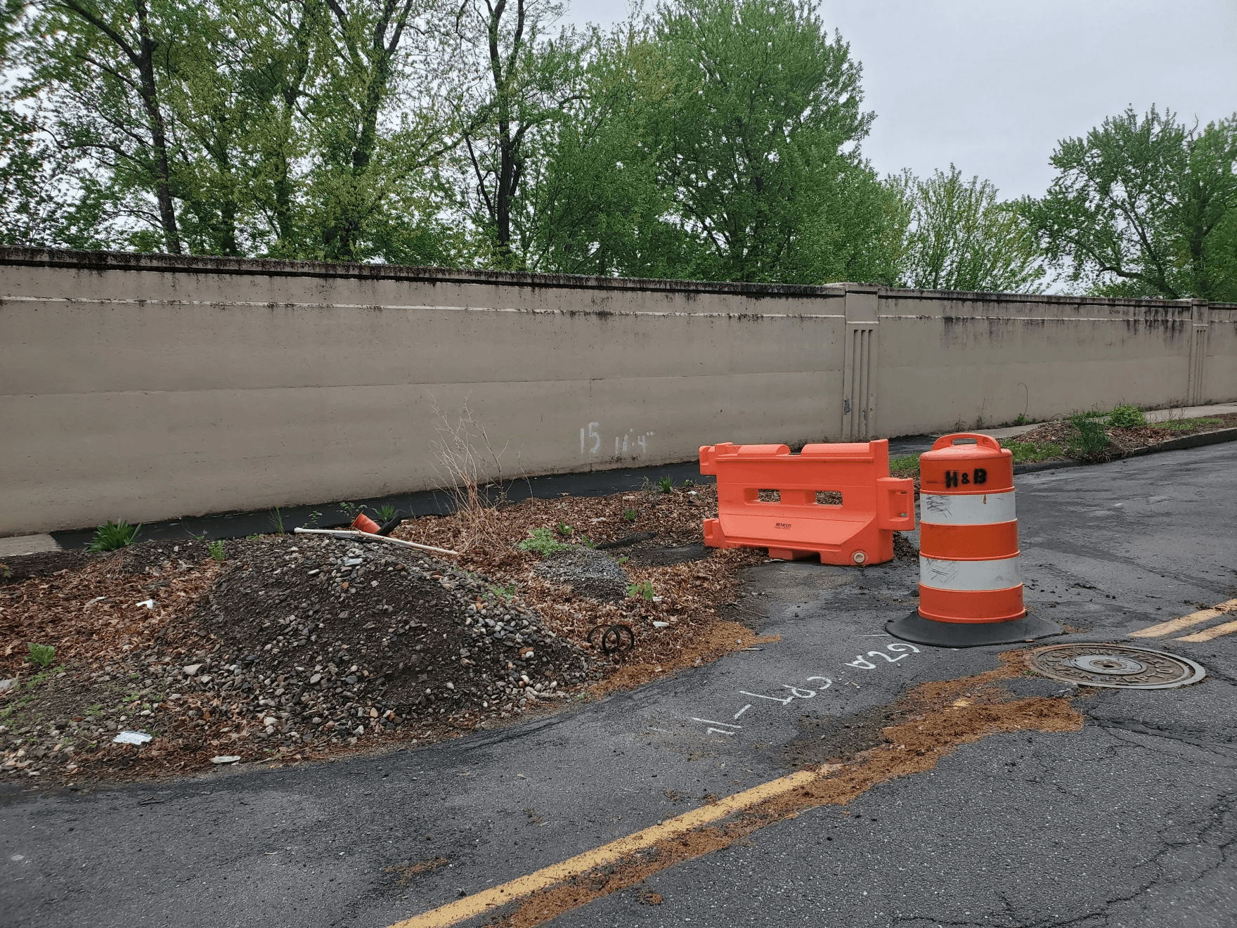 A patched sinkhole on Riverside Road in Springfield, Massachusetts, filled with dirt and rocks. An orange traffic barrel and barrier sit beside a cracked roadway near a flood protection wall.