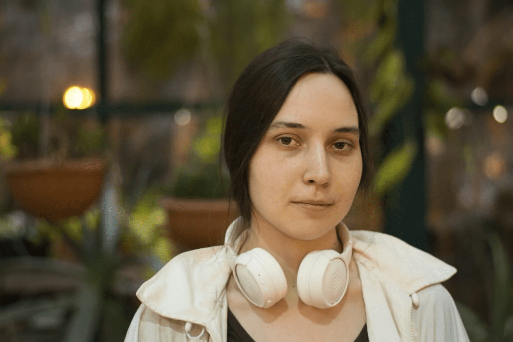 Kseniia Petrova, a young woman with long dark hair, wearing a light-colored jacket and white headphones around her neck, stands in a garden-like setting with blurred plants in the background.