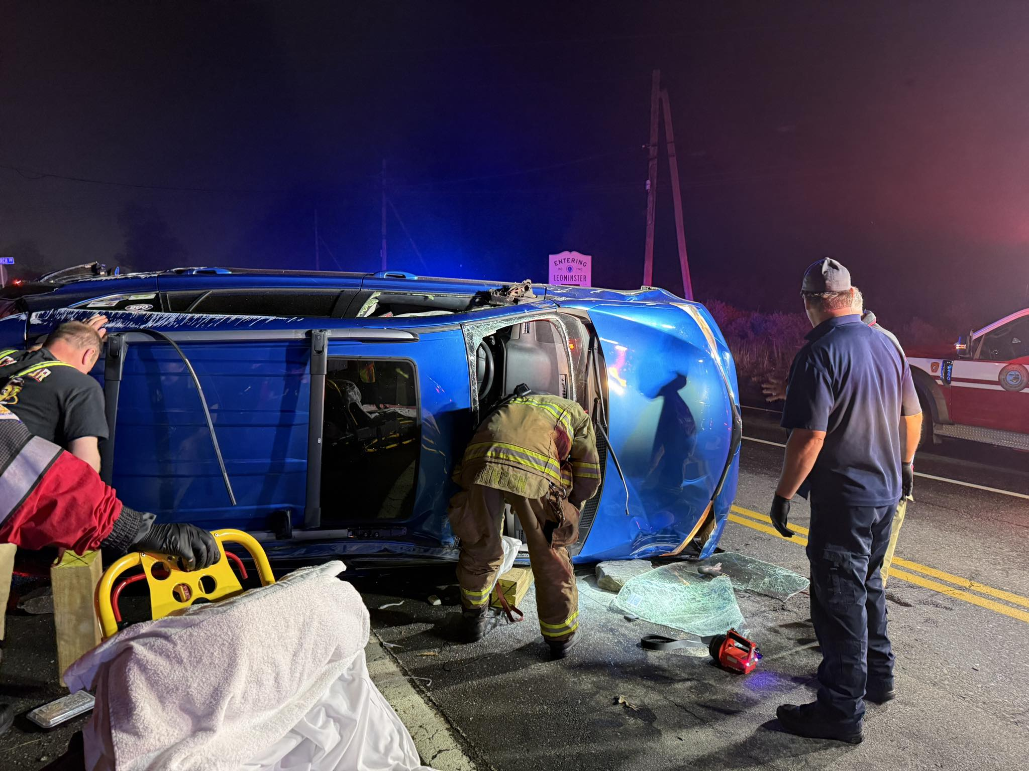 Firefighters respond to a rollover crash at night involving a blue SUV lying on its side on Leominster Road near a sign that reads “Entering Leominster.” Emergency personnel use rescue tools next to broken glass.