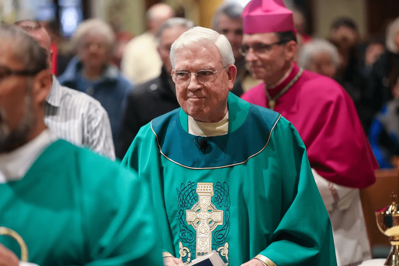 Monsignor Francis Strahan, wearing green liturgical vestments with a gold-embroidered cross, walks down the aisle of a church during a service. A bishop in pink vestments and congregants are visible in the background.