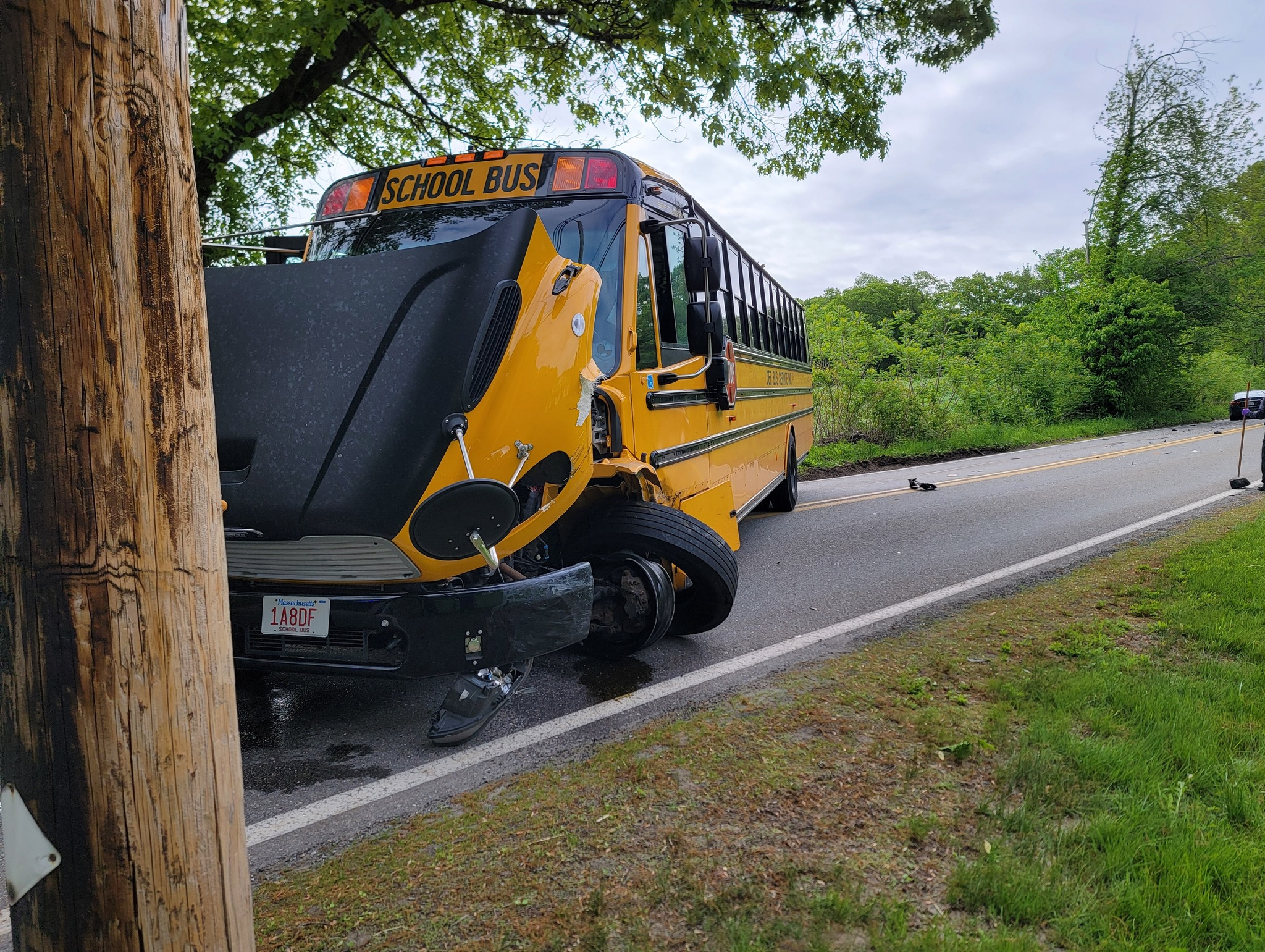 A damaged yellow school bus with a crumpled front end is parked on the side of a rural road near a utility pole. Debris is scattered on the pavement. The photo was taken by the Lunenburg Police Department.