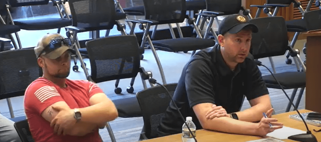 Two men sit with arms crossed at a public meeting table during the April 29, 2025 Fitchburg Public Works Committee meeting. One wears a red shirt and sunglasses; the other wears a black polo and baseball cap with a yellow patch, speaking into a microphone.