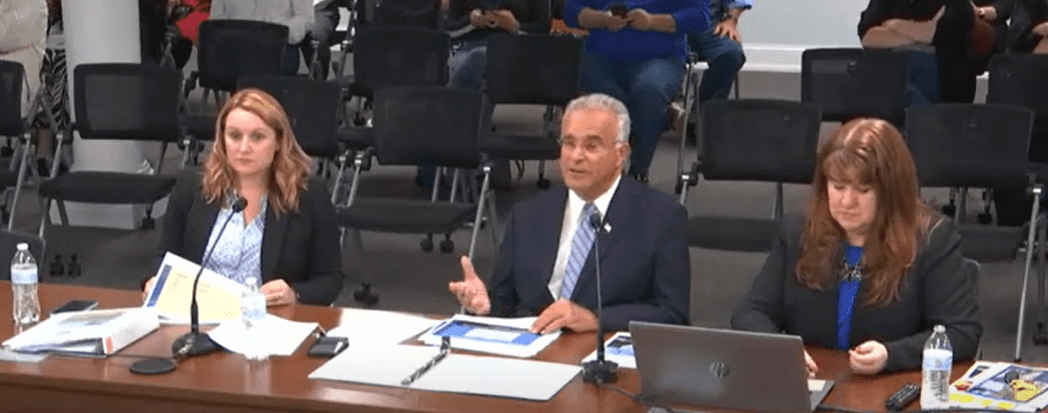 Leominster Mayor Dean Mazzarella, seated between two city officials, speaks during a Finance Committee meeting. Papers, microphones, and laptops are on the table in front of them. The meeting took place on May 19, 2025. Photo by Leominster Access Television.