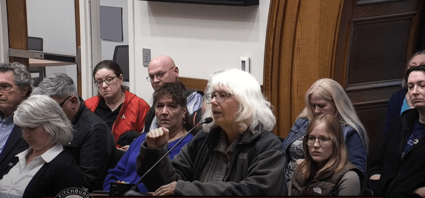 A woman with white hair and glasses speaks into a microphone at a public meeting, addressing the Fitchburg City Council. Seated attendees behind her listen intently, some appearing serious or concerned.