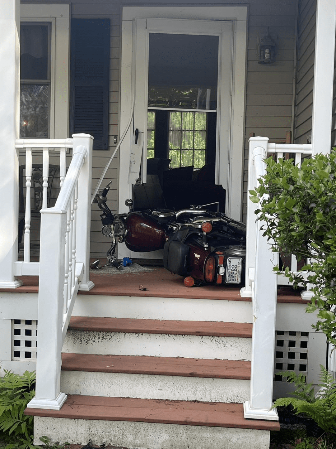 A red motorcycle is seen tipped over on the front porch of a beige house, having crashed into the front door. The storm door is cracked and partially open, and the scene shows minor structural damage.