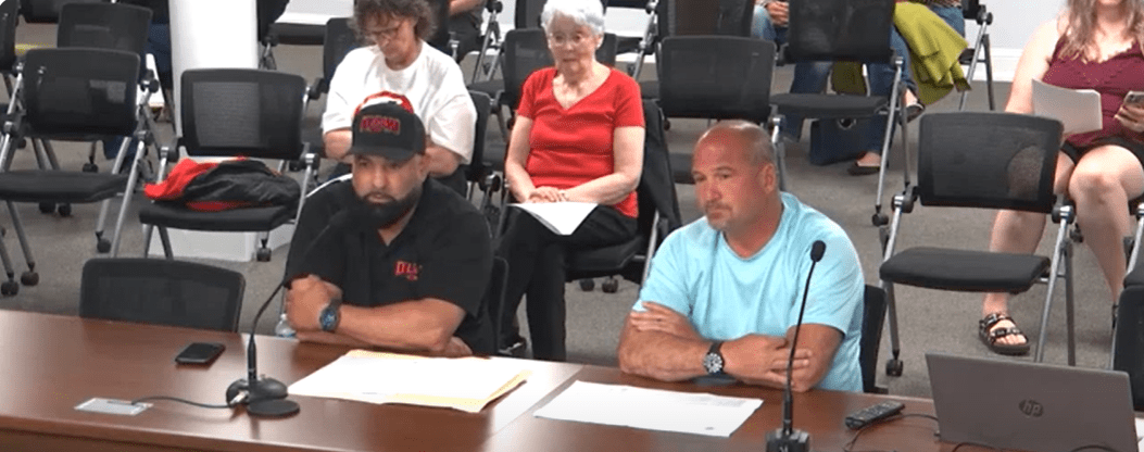 Two men sit at a table speaking into microphones during a city council meeting. The man on the left wears a black shirt and hat with “DSF” on it; the man on the right wears a light blue shirt. Papers and microphones are in front of them, and members of the public are seated behind them.
