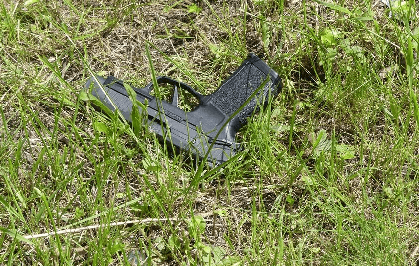 A black handgun lying in tall grass, partially concealed by weeds. The firearm appears to be abandoned in an outdoor setting.