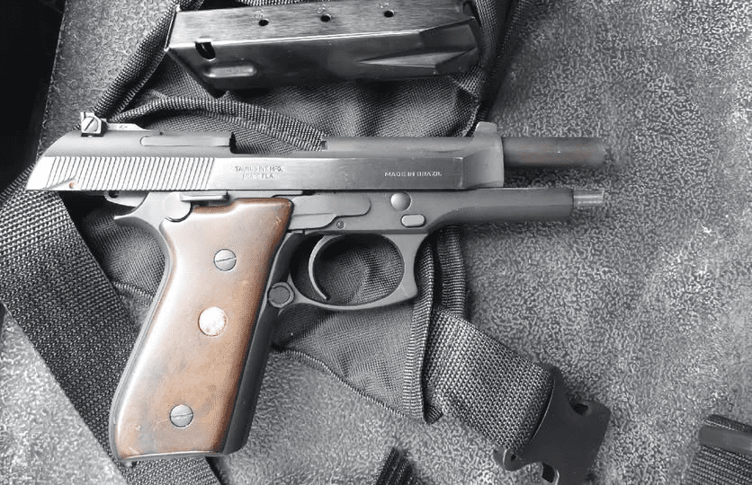 A black semi-automatic handgun with a wooden grip, partially disassembled with the slide back and magazine removed, resting on a black tactical bag.