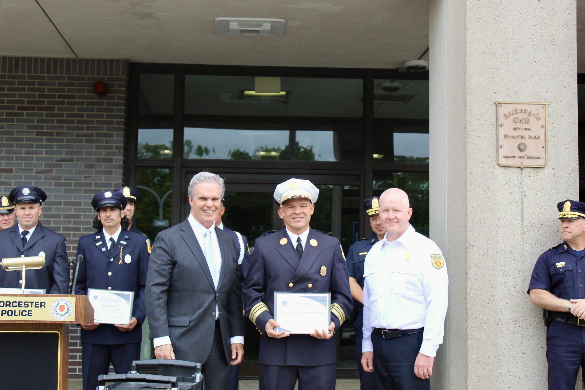 A Worcester firefighter in full dress uniform holds a certificate while posing with DA Joseph D. Early Jr. and Fire Chief Martin Dyer in front of the Worcester Police Department.