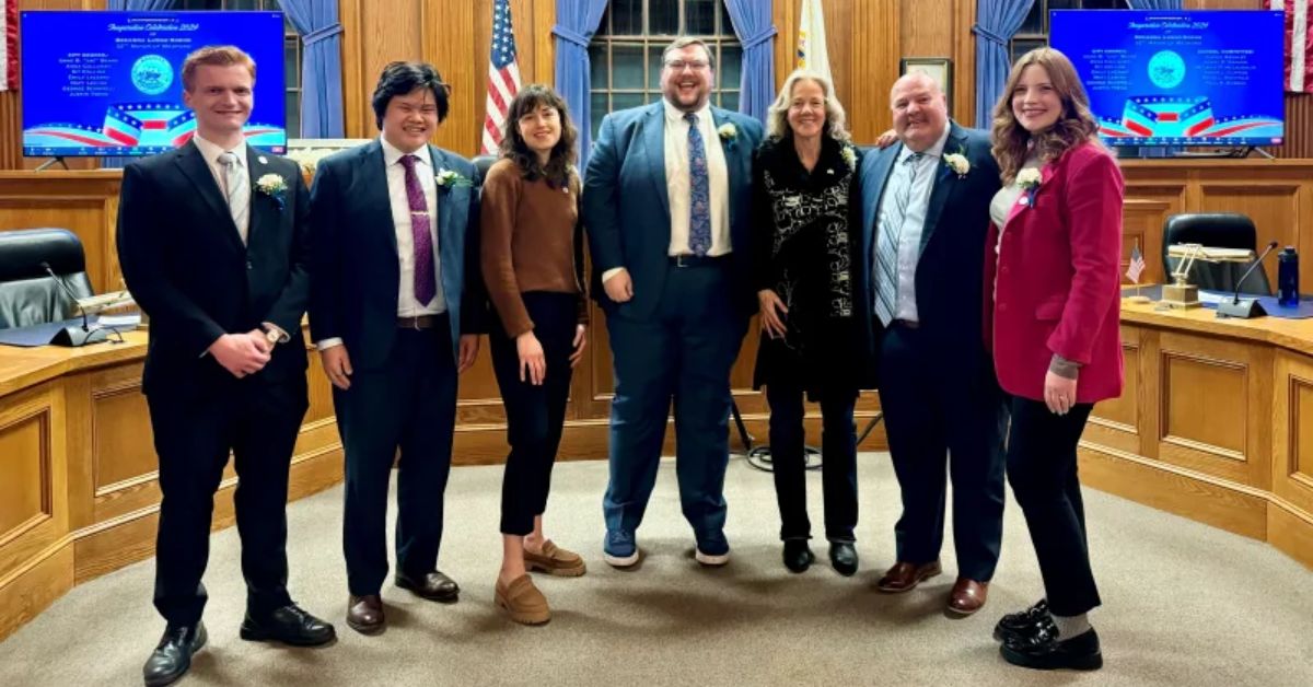 Seven Medford City Council members stand side by side in the council chamber, smiling at the camera. The chamber features wooden paneling, American and Massachusetts flags, and screens displaying the city seal.