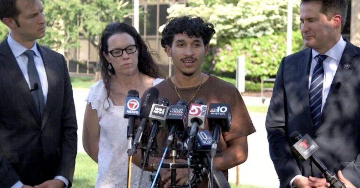 Marcelo Gomes da Silva, an 18-year-old in a brown shirt, stands at a podium surrounded by microphones, speaking to the press. To his left and right are U.S. Representatives Jake Auchincloss and Seth Moulton, and attorney Robin Nice. The group stands outdoors with trees and a building in the background.