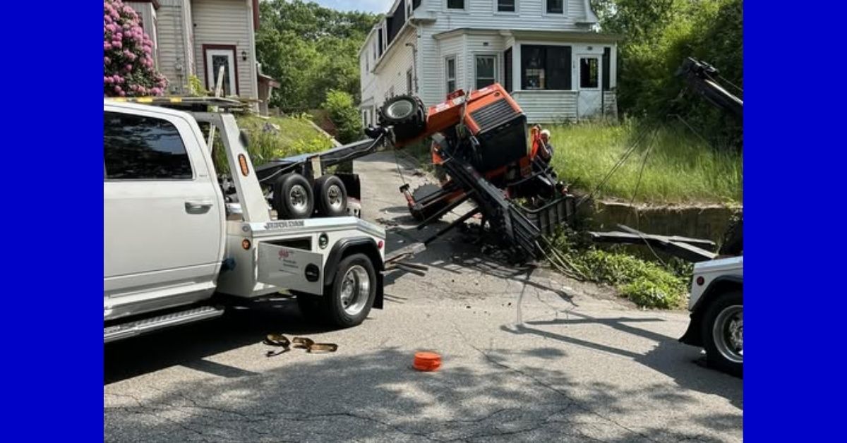 Overturned forklift on a steep driveway in front of a white house, with two tow trucks on scene in Athol following a delivery accident.