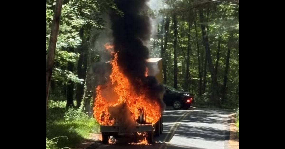 A box truck fully engulfed in flames on a wooded roadway, sending thick black smoke into the sky as it burns.