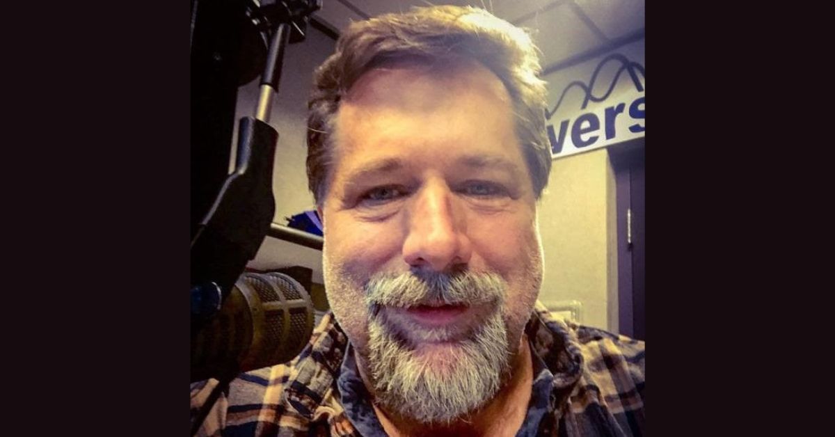 George Knight, a middle-aged man with a gray beard and mustache, smiles at the camera in a radio studio with a microphone visible beside him.