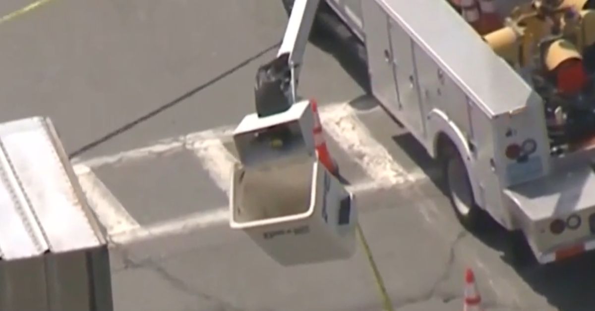 Aerial view of a white utility bucket truck positioned in an intersection, with the bucket extended and empty. Traffic cones and part of a large trailer truck are also visible.